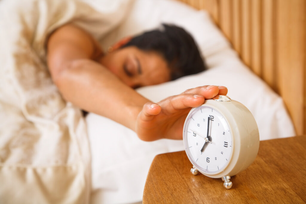 Bild einer Frau mit kurzen dunklen Haaren liegt im Bett und stellt einen weißen Wecker aus.