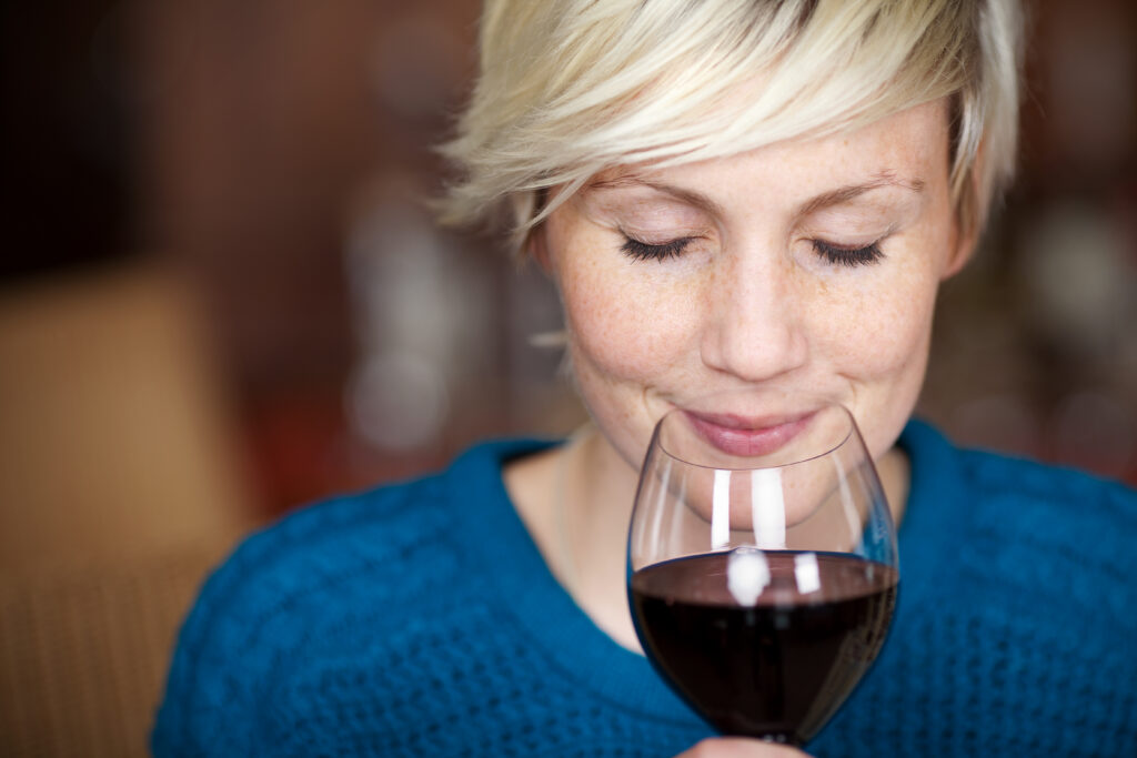 Bild einer jungen Frau, die mit geschlossenen Augen an einem Glas Wein riecht und lächelt.