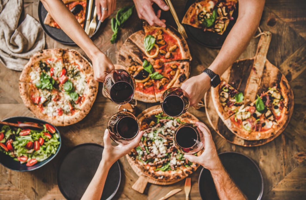 Bild von vier Personen am Tisch mit verschiedenen Pizzen stoßen mit Wein an.